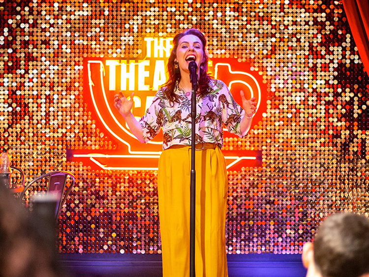A woman singing in front of a gold sequin wall on stage.